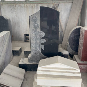 "Black granite headstone with carved sunflower relief on left side, photographed in our factory among other stone components and tools."