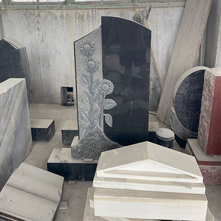 "Black granite headstone with carved sunflower relief on left side, photographed in our factory among other stone components and tools."