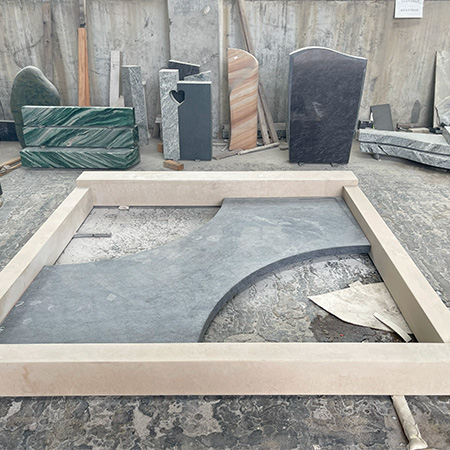 "Custom granite ledger framed by light-colored stone with curved dark centerpiece, photographed in ELHASG workshop among upright headstones and sculptural pieces."