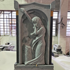 "Black granite memorial sculpture of a woman in flowing robes within an arched alcove, photographed inside ELHASG factory workshop with tiled walls and stone blocks."