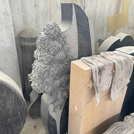 "Hand-carved headstone memorial sculpture featuring a deer beneath a layered tree, photographed in workshop with tools and materials."