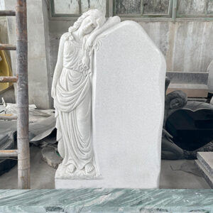 "Unfinished marble memorial headstone with sculpted angel resting peacefully on blank panel, photographed in our factory surrounded by tools and stone components."