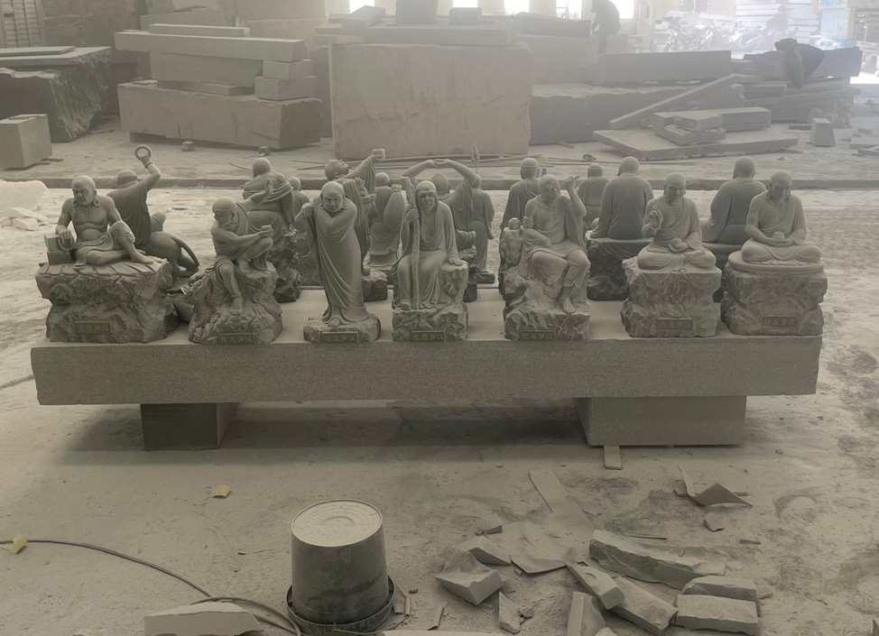 "Seated stone Arhat statues in Elhasg workshop with inscriptions and tools in background."