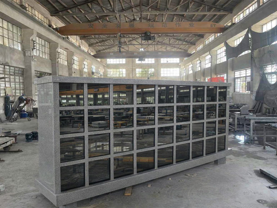 "Grey granite columbarium featuring black niche doors inside a stone fabrication workshop with overhead crane."